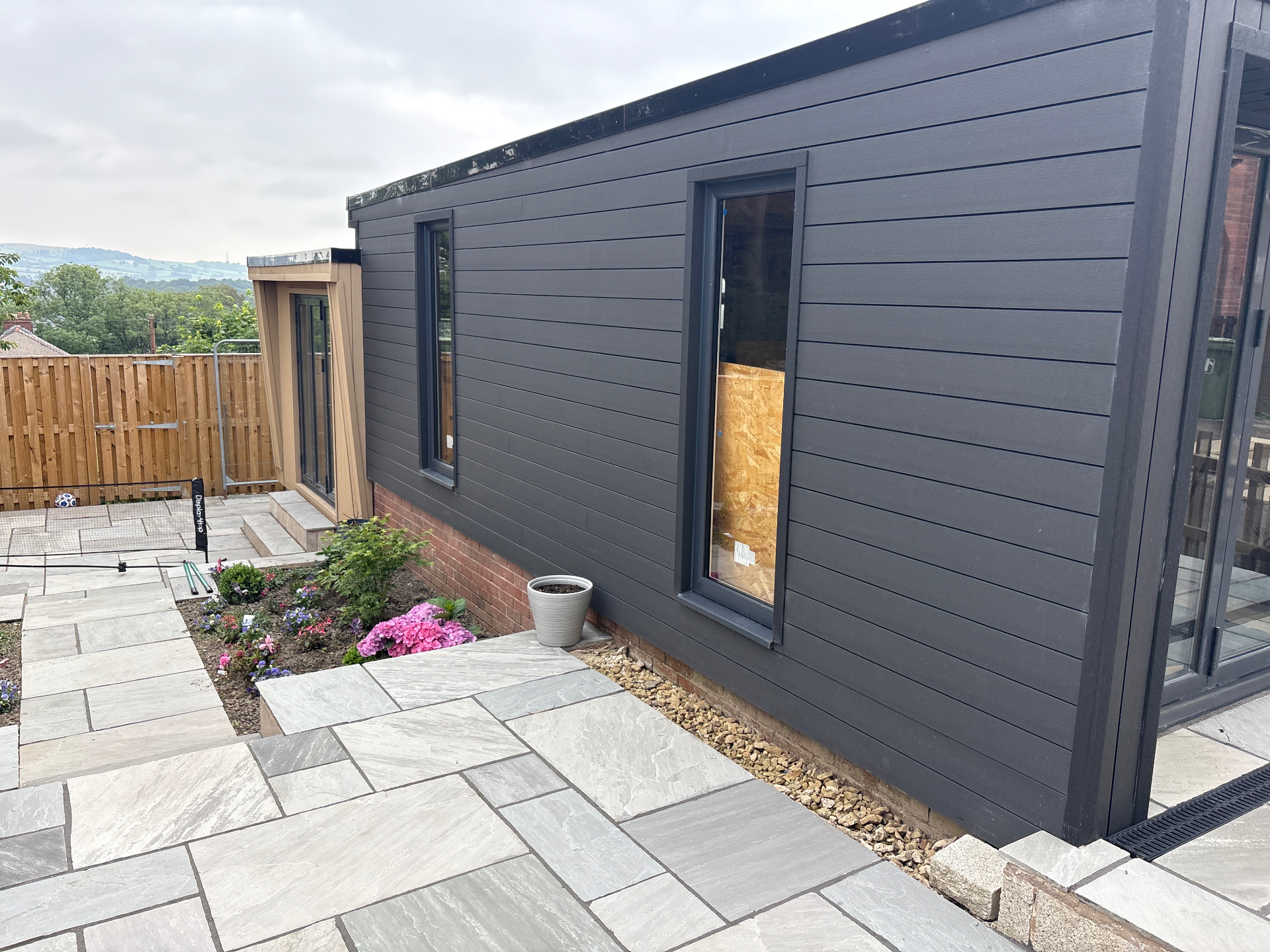 Side elevation — flush composite boards with portrait windows, gravel French drain and brick plinth.